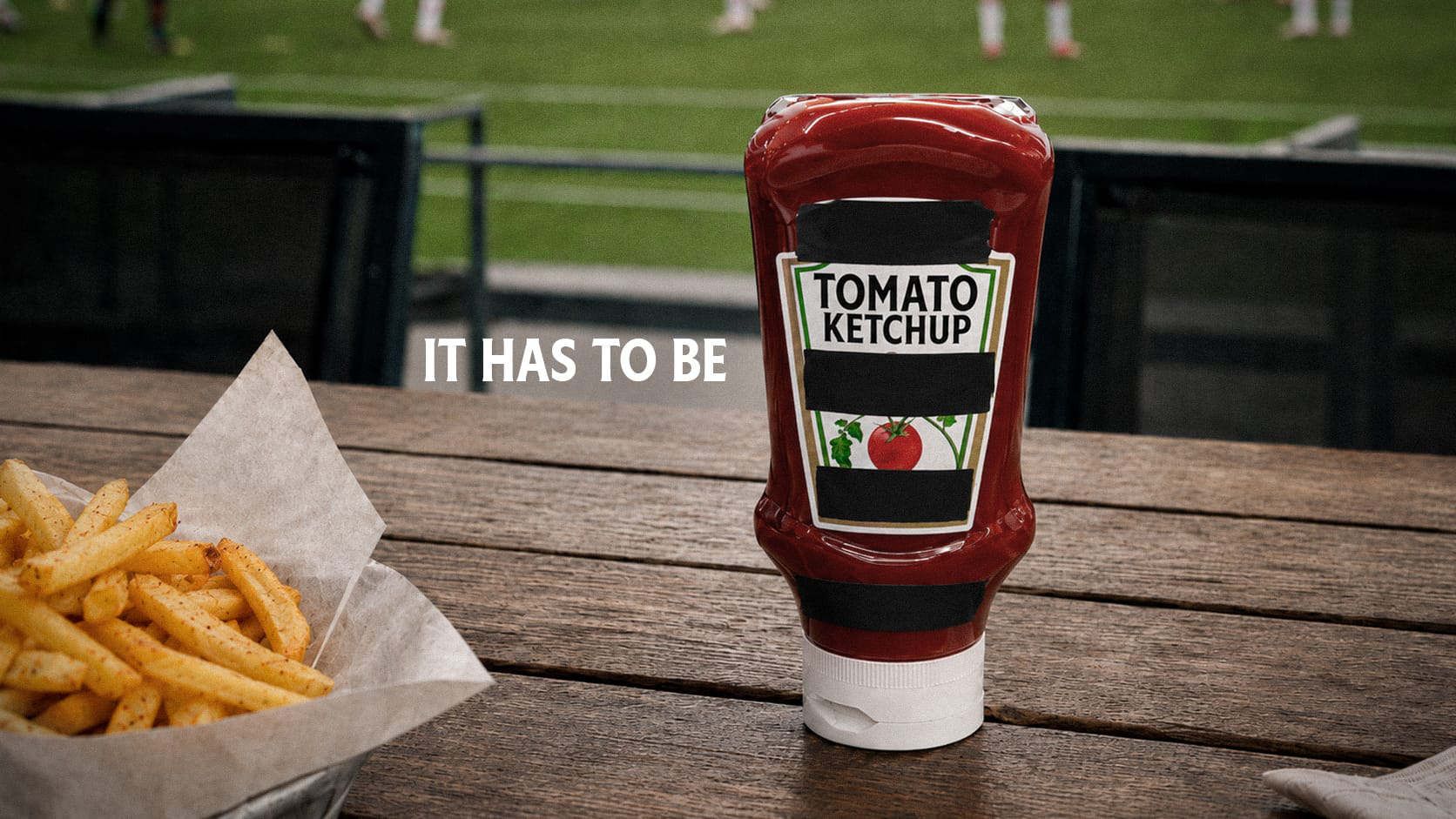 Photographie d'un packaging de ketchup Heinz avec un scotch noir sur la marque, des frites à côté, on devine un match de foot en fond